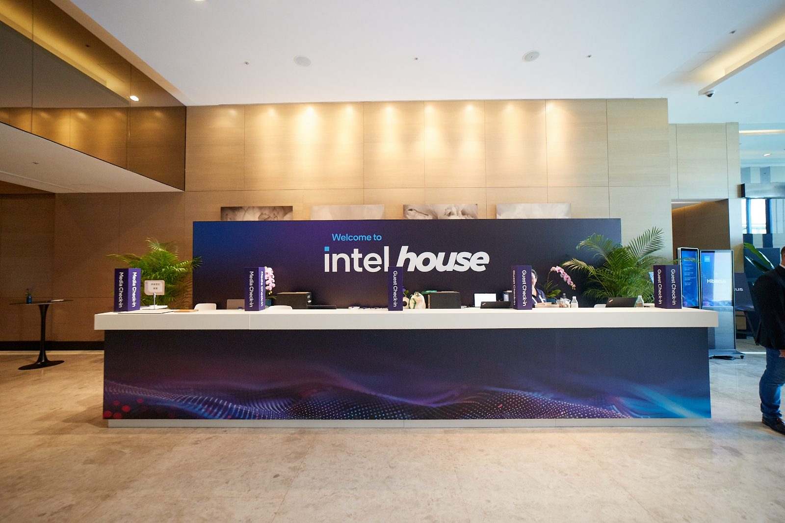 The Intel House check-in: a long white counter with media and guest check-in paddles in front of a Welcome to Intel House graphic wall with orchids and palms
