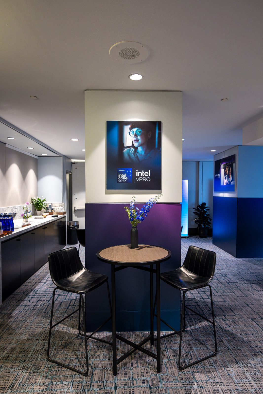 A pre-function vignette with a backlit Core Ultra Series 3 and vPro poster, cafe seating, and delphinium in a bud vase
