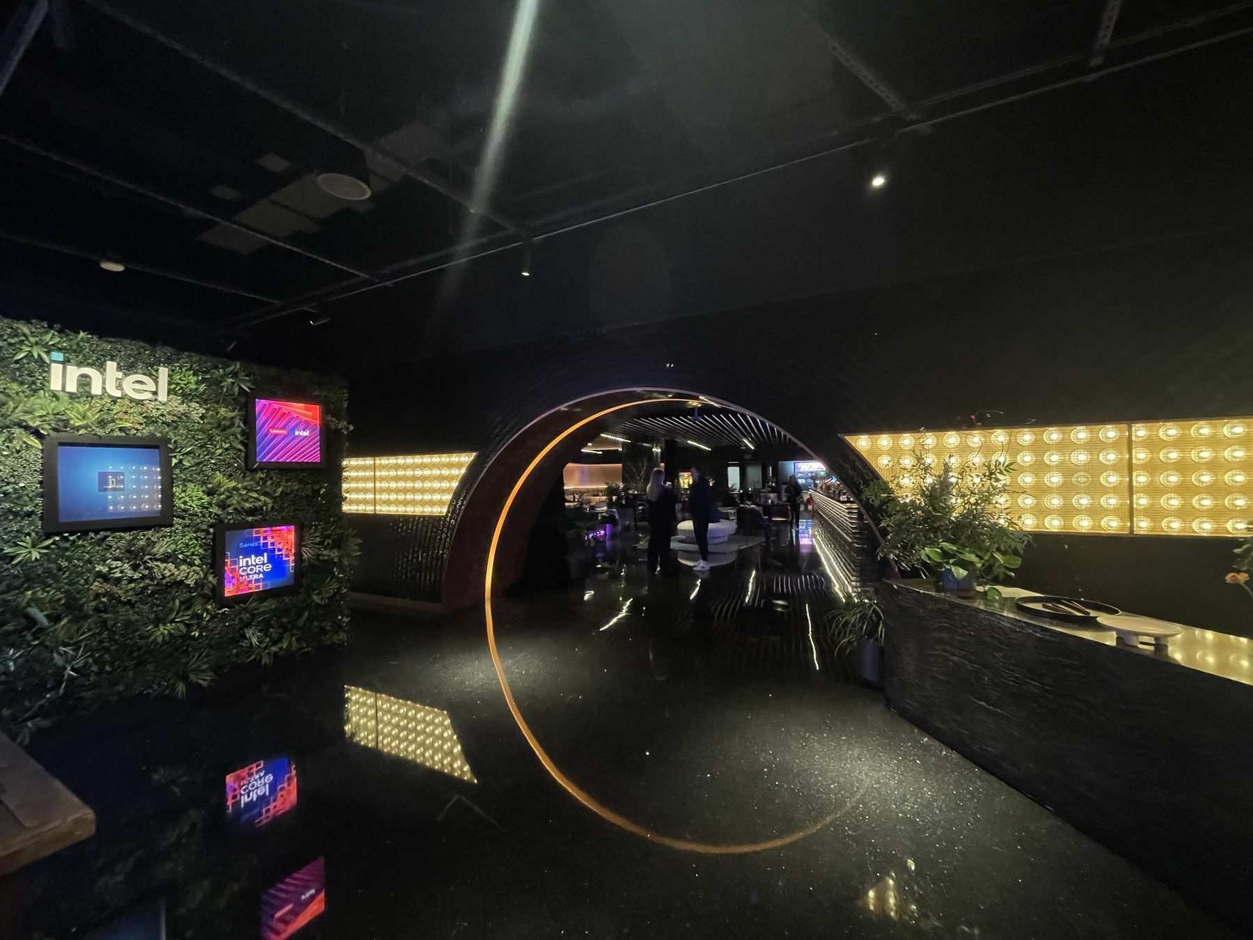 Entrance to the Intel Oasis: a living green wall with the white Intel logo and embedded LED screens, facing a circular arch rimmed in warm orange light that opens into the lounge