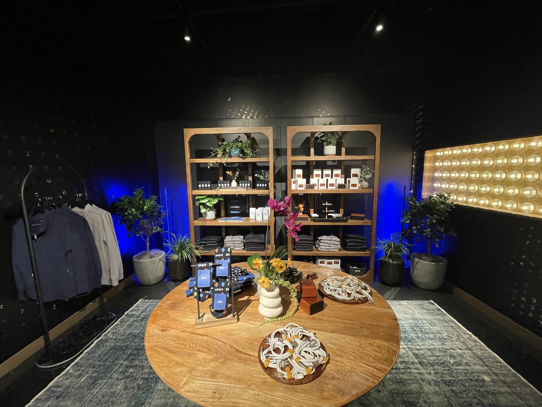 Marketplace vignette with a round wood table of Intel-branded merch and florals, tall arched wood shelving behind it, apparel rack to the left, and plants throughout